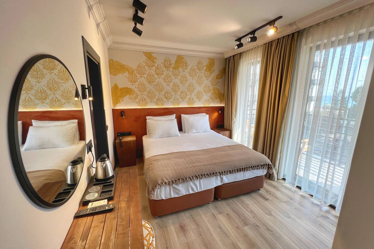 A hotel room with a large bed, a bedside table with a lamp, a mirror, and a view of the outdoors through large windows.