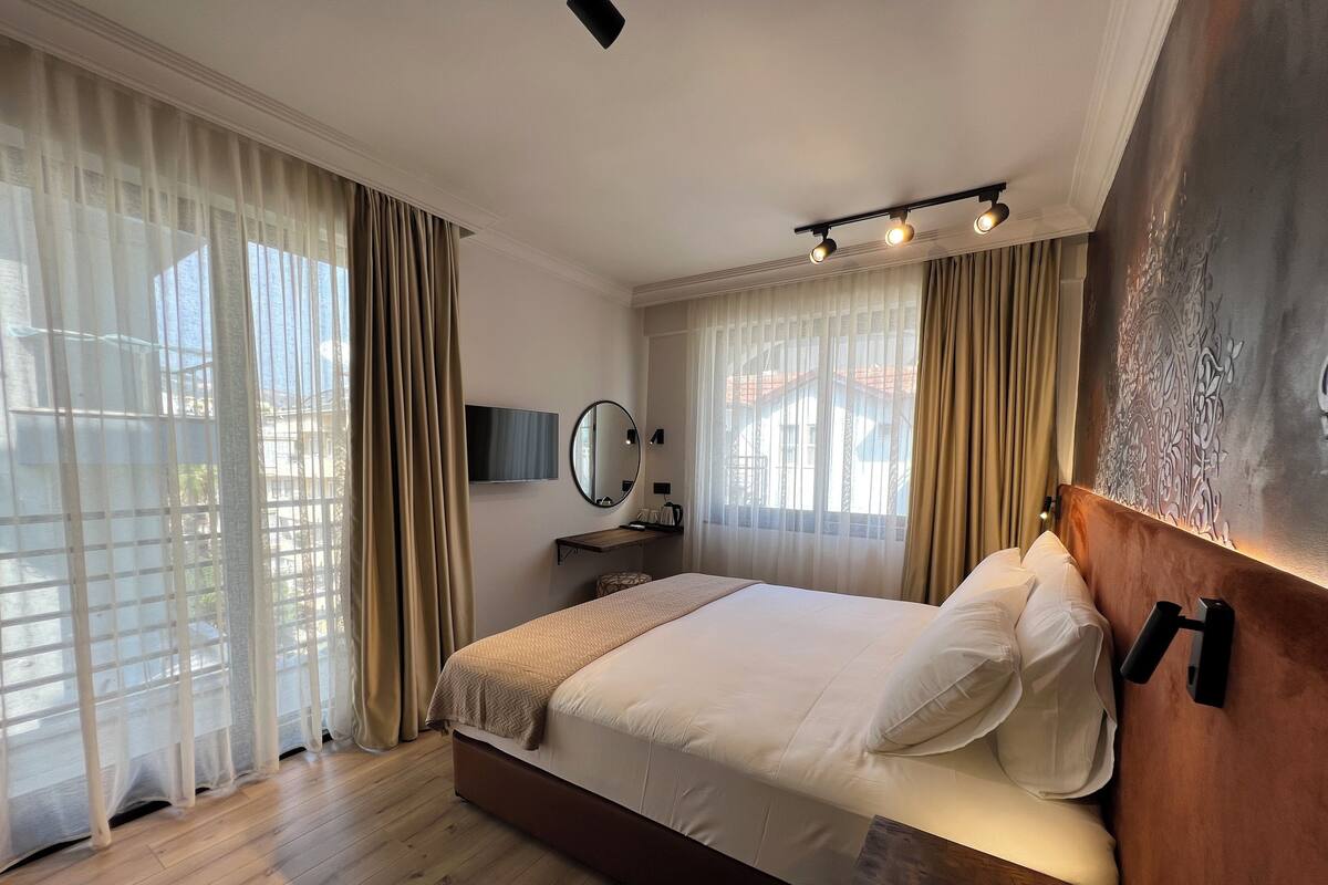 A modern hotel room with a large bed, a bedside table, a TV, and a window with curtains.