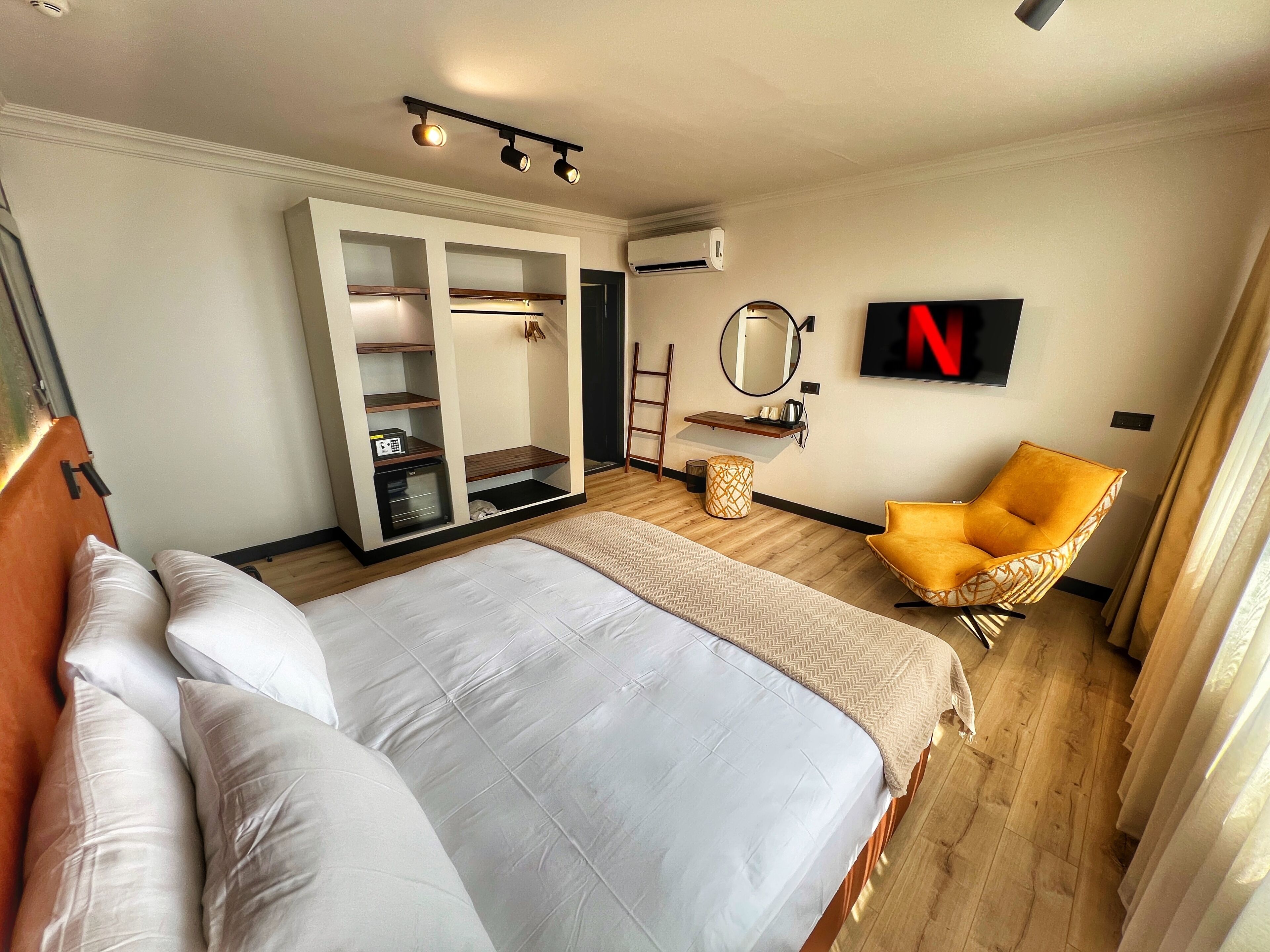 Una habitación de hotel moderna con una cama grande, un armario con estantes, un espejo redondo pequeño, un sillón amarillo y un televisor que muestra el logotipo de Netflix.