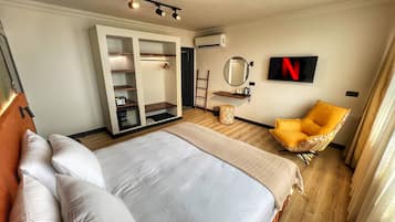 Una habitación de hotel moderna con una cama grande, un armario con estantes, un espejo redondo pequeño, un sillón amarillo y un televisor que muestra el logotipo de Netflix.