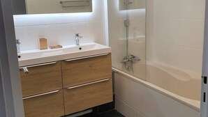 Un baño moderno con un tocador blanco, cajones de madera, una cabina de ducha con paredes de vidrio y un espejo grande.