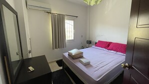 A bedroom with a bed, a bedside table, a lamp, a window with curtains, and an air conditioning unit. - 98. Homey 2BR Townhouse | 2 Queen Beds | Garden (Sheikh Zayed City)