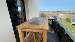 Une terrasse en bois avec une table et des chaises, donnant sur une zone herbeuse et offrant une vue sur l’océan et les éoliennes.