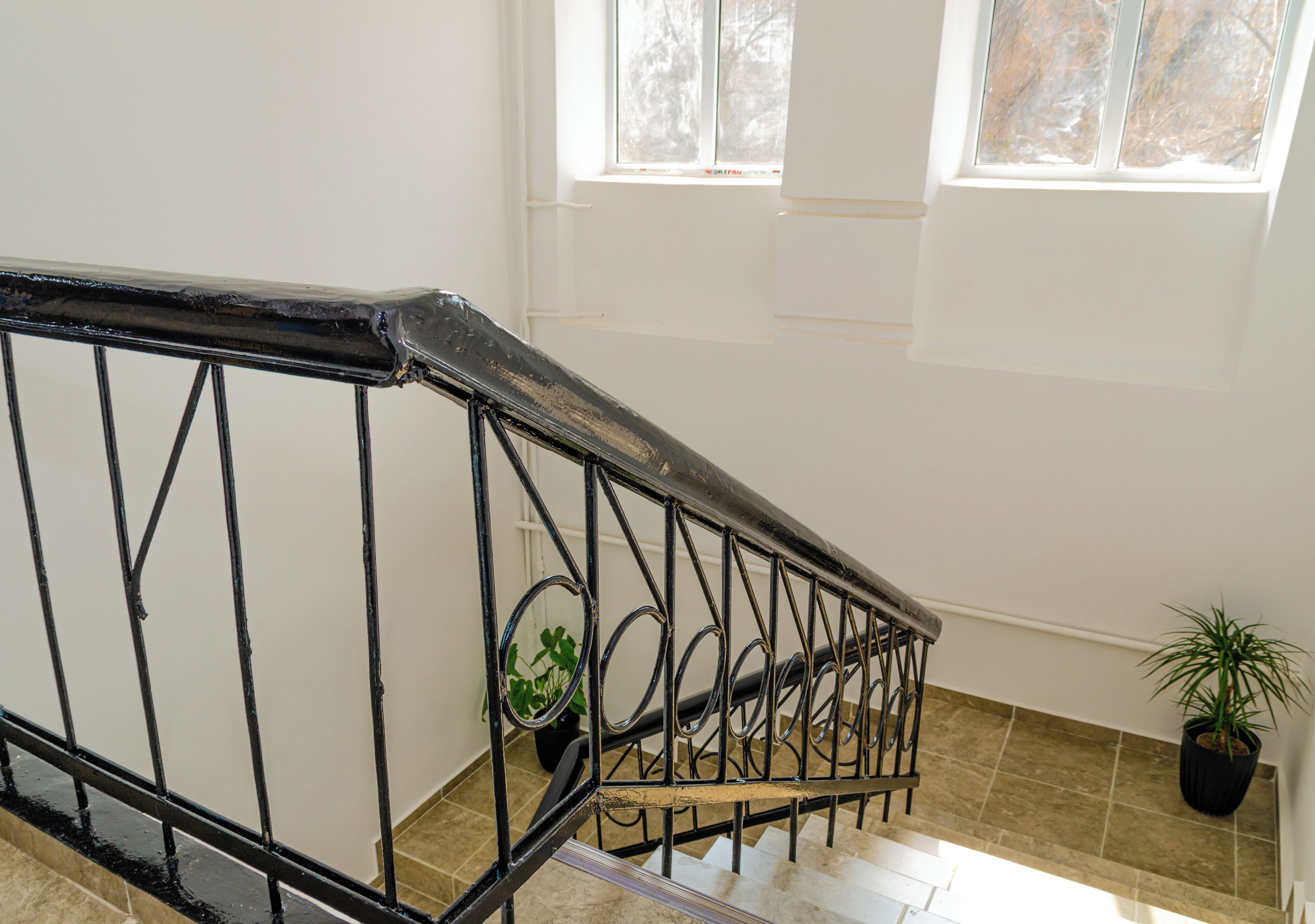 Eine Treppe mit schwarzem Metallgeländer, Pflanzkübeln und Blick auf Bäume durch ein Fenster.