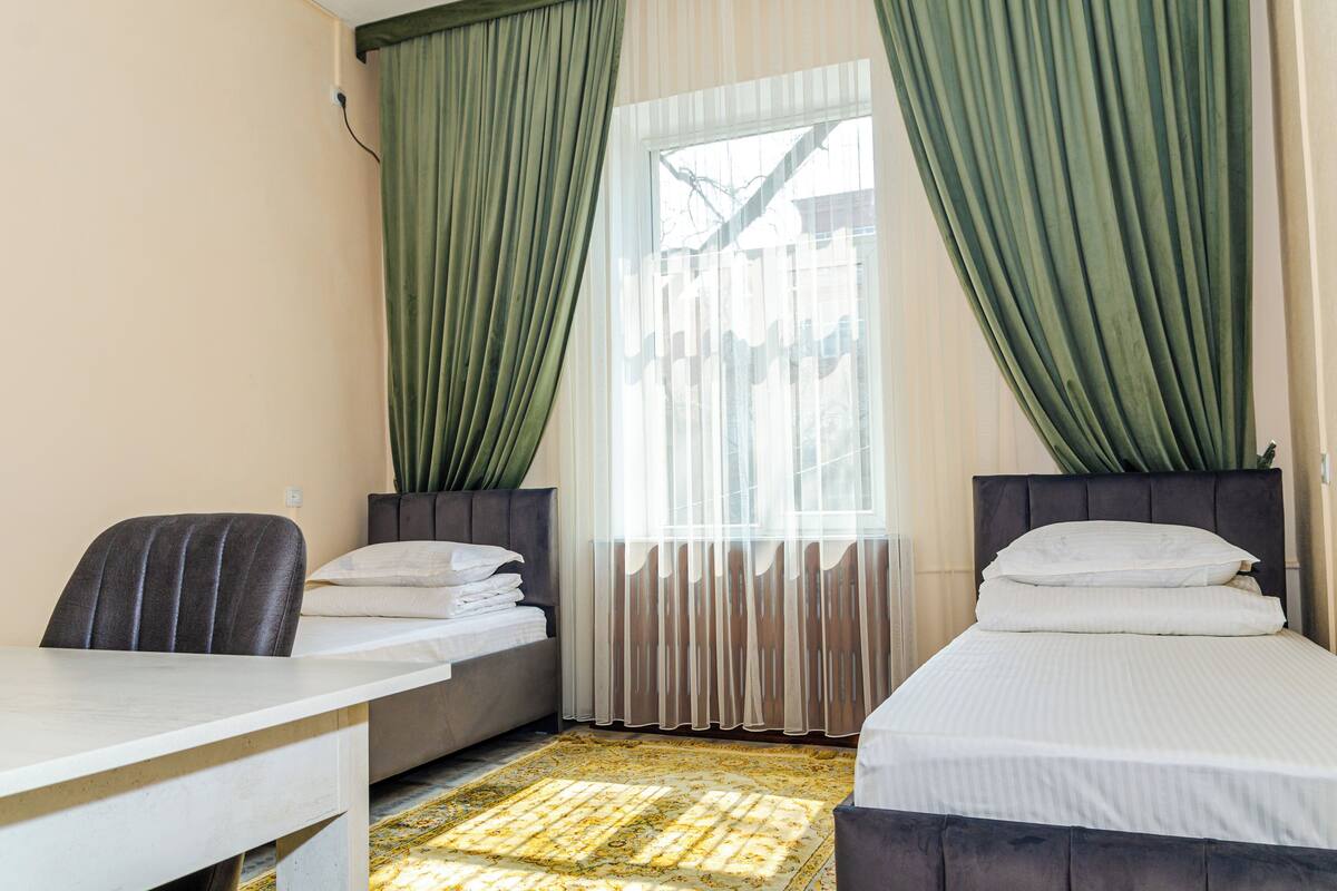 A hotel room with two beds, a desk, and a window with sheer curtains.
