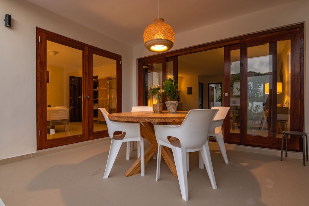 A dining area with a wooden table and white chairs, a hanging light fixture, and a view through glass doors.