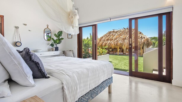 A bedroom with a large bed, a bedside table, a mirror, and a view of a thatched-roof structure outside. - Villa Blou, steps from Jan Thiel Beach (Jan Thiel)