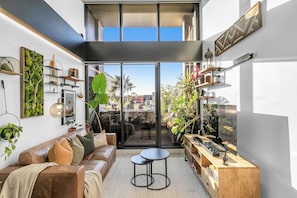 A modern living room with a leather sofa, two round tables, and a large window overlooking a cityscape. - Sol Docklands | Large 3BR Loft, Infinity Pool, Gym, Free Parking & Free Trams (Docklands)