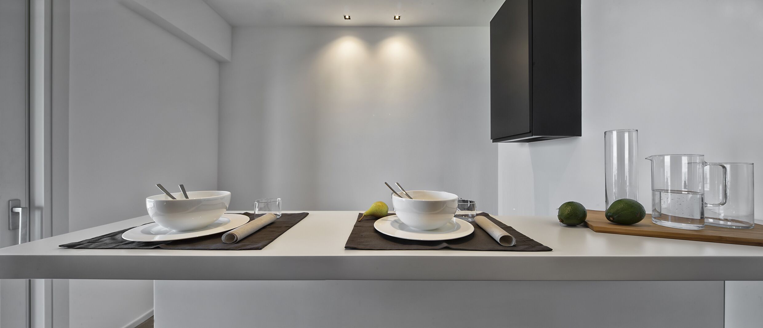 Una cucina moderna con ripiani bianchi, due ciotole bianche con cucchiai, una pera, due frutti verdi e bicchieri trasparenti.