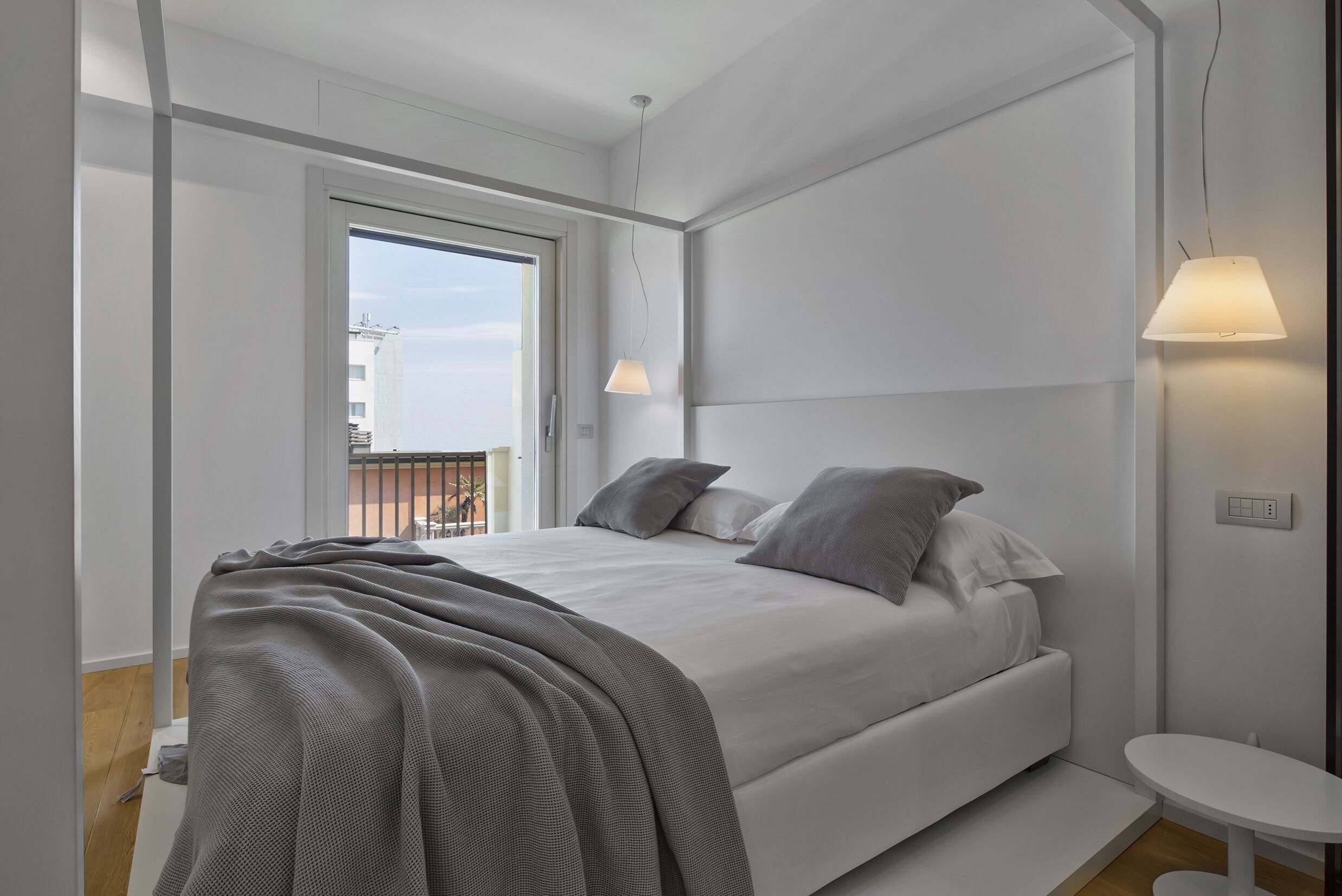 A modern bedroom with a large bed, a small bedside table, and a window with a view of buildings.