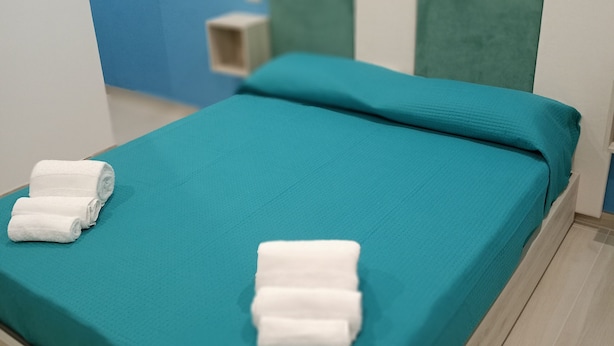A neatly made bed with teal bedding and two rolled white towels, a wooden headboard, and a blue wall with a small shelf.