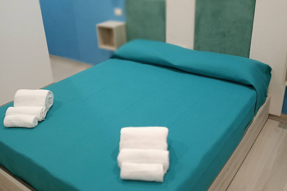 A neatly made bed with teal bedding and two rolled white towels, a wooden headboard, and a blue wall with a small shelf.