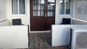 Une entrée de maison avec une porte marron, deux fenêtres à cadres blancs et un appareil de climatisation blanc sur le côté droit.