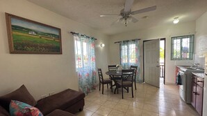 Un salon avec un canapé marron, une table à manger avec des chaises, un ventilateur de plafond et un coin cuisine avec une cuisinière.