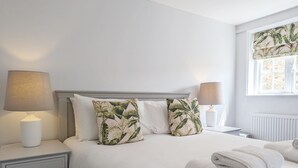 Ein ordentlich bezogenes Bett mit weißen Leinen, zwei Zierkissen mit tropischem Blattmuster, eine Nachttischlampe und ein aufgerolltes weißes Handtuch auf dem Bett.