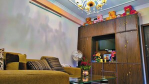 Un salon comprenant un canapé, une table basse en verre, un meuble en bois orné d’ours en peluche, un ventilateur et un lustre.