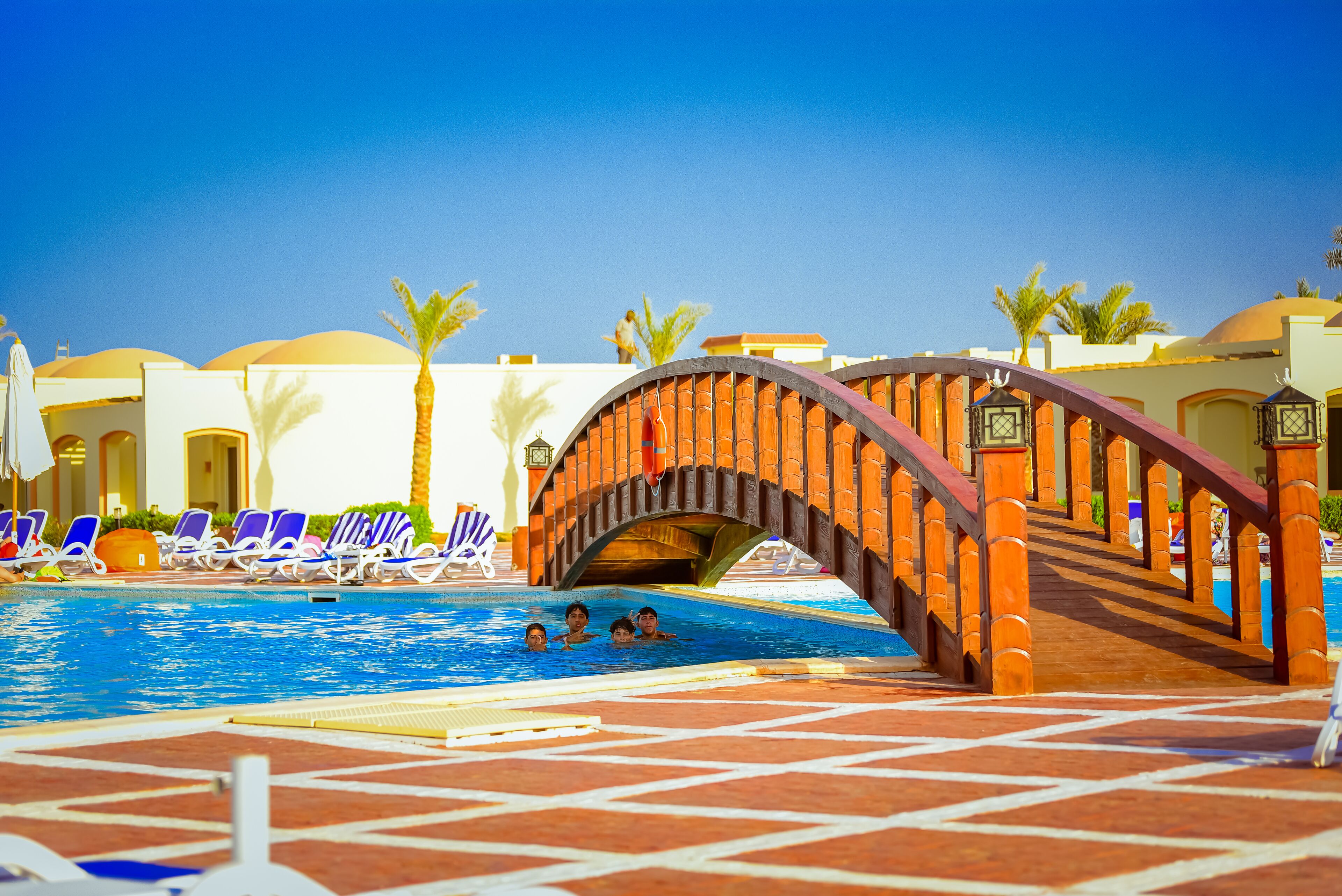 A pool area with a brick arch bridge, lounge chairs, and palm trees.