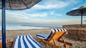 A beachside lounge with striped towels and wooden lounge chairs, a thatched umbrella, and a pier in the background. - Amarina Queen Resort & Aqua Park Marsa Alam (Marsa Alam)