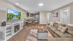 Una sala de estar con chimenea, televisor, comedor y cocina al fondo.