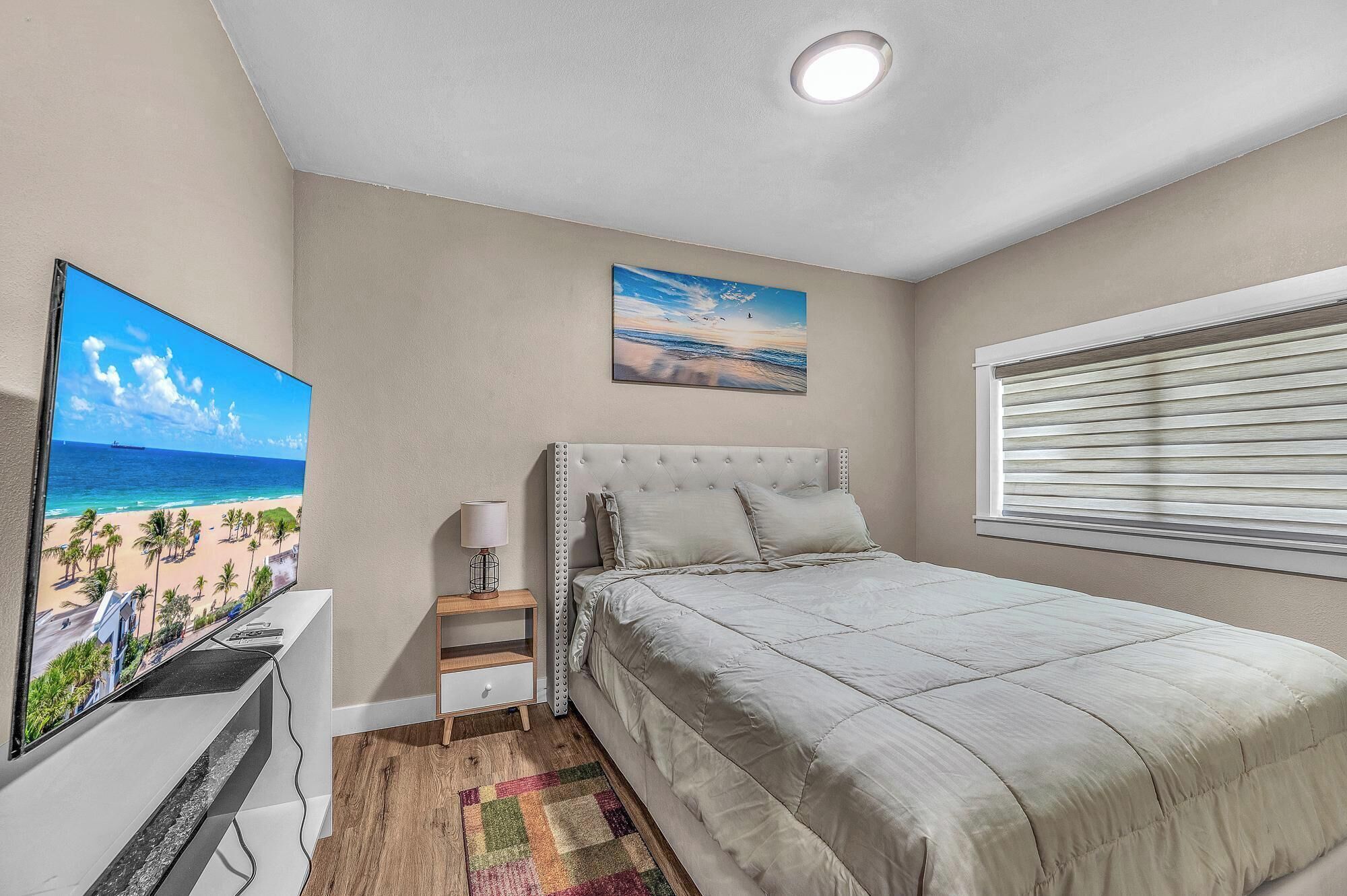 Camera da letto con un letto grande, un comodino, una lampada, una televisione e un quadro raffigurante una spiaggia.