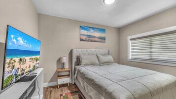 Camera da letto con un letto grande, un comodino, una lampada, una televisione e un quadro raffigurante una spiaggia.