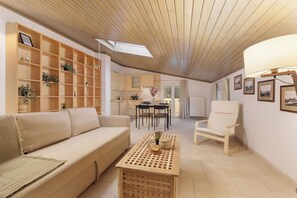 A spacious living area with a wooden ceiling, a sofa, a coffee table, and a chair. There is a kitchen area with cabinets and a dining table. - Country Attic (Corfu)