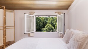 Un dormitorio con una cama grande, una ventana con cortinas abiertas y vistas a árboles desde afuera.