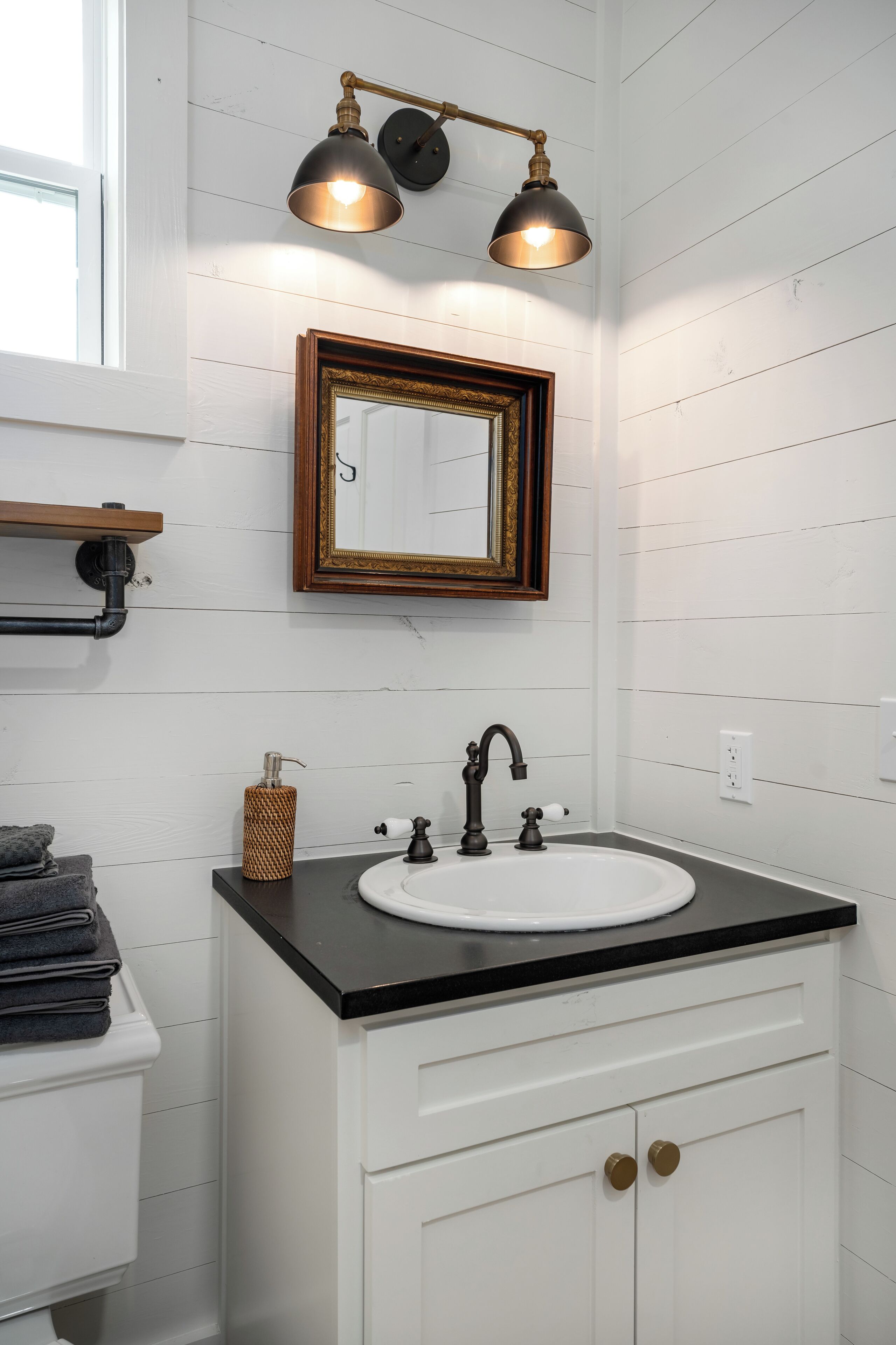 A bathroom with a white vanity, a black countertop, a white sink, a gold faucet, a mirror with a gold frame, and a wall-mounted light fixture.