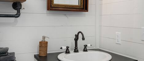 A bathroom with a white vanity, a black countertop, a white sink, a gold faucet, a mirror with a gold frame, and a wall-mounted light fixture.