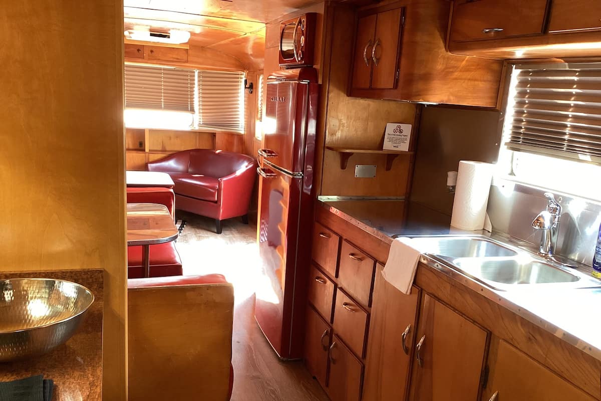 A compact RV kitchen with wooden cabinets, a refrigerator, a sink, and a dining area with a red couch.