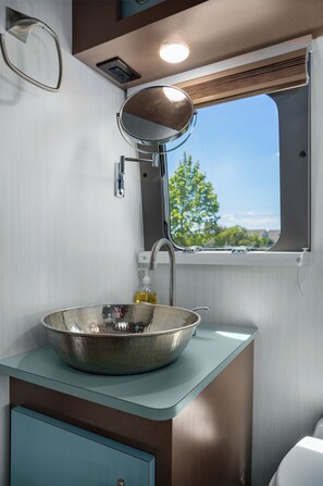 A modern bathroom with a large mirror, a stainless steel sink, and a window offering a view of trees and a road.