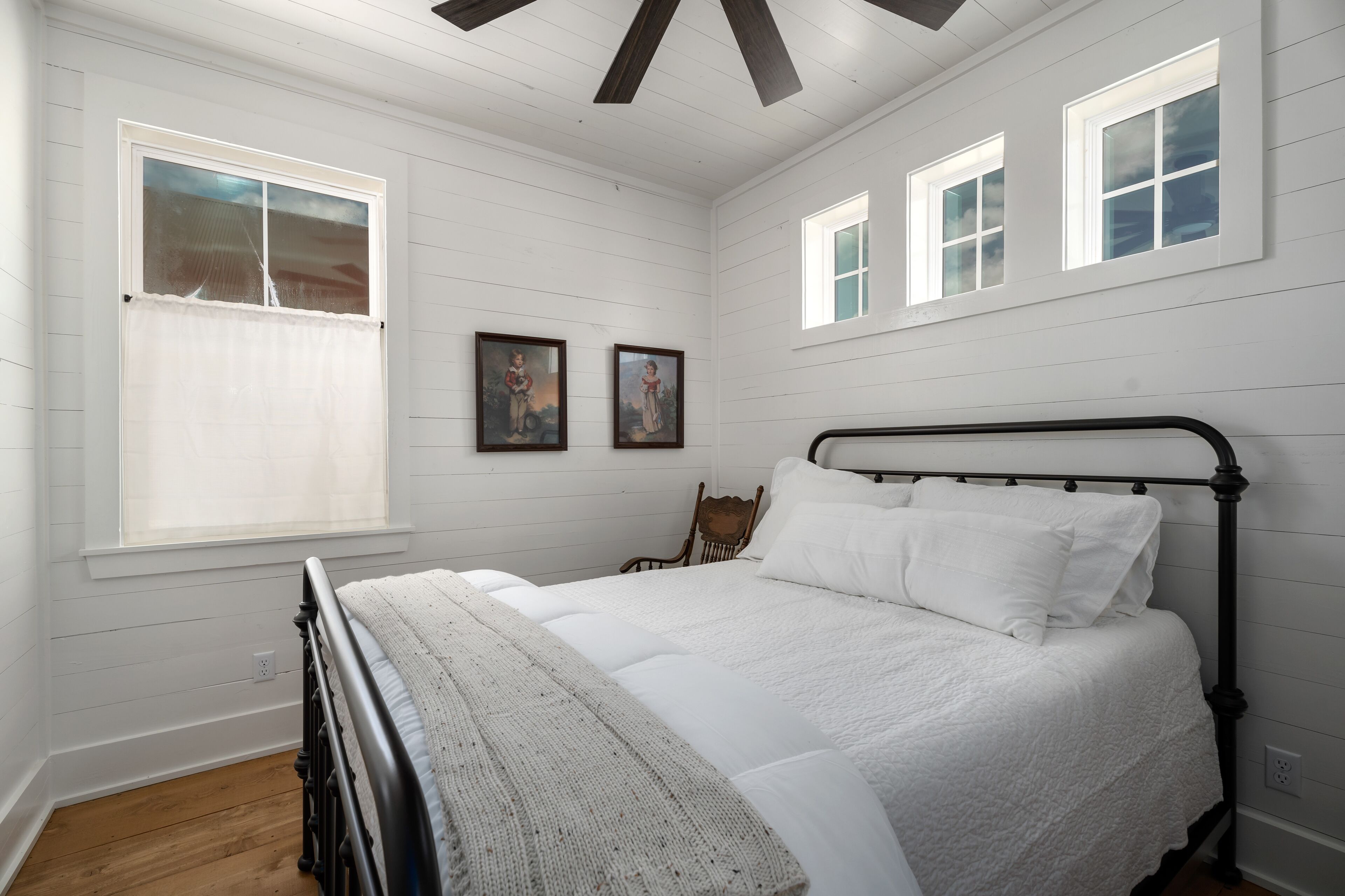 Une chambre avec un grand lit, un plancher en bois, des murs blancs et un ventilateur de plafond.