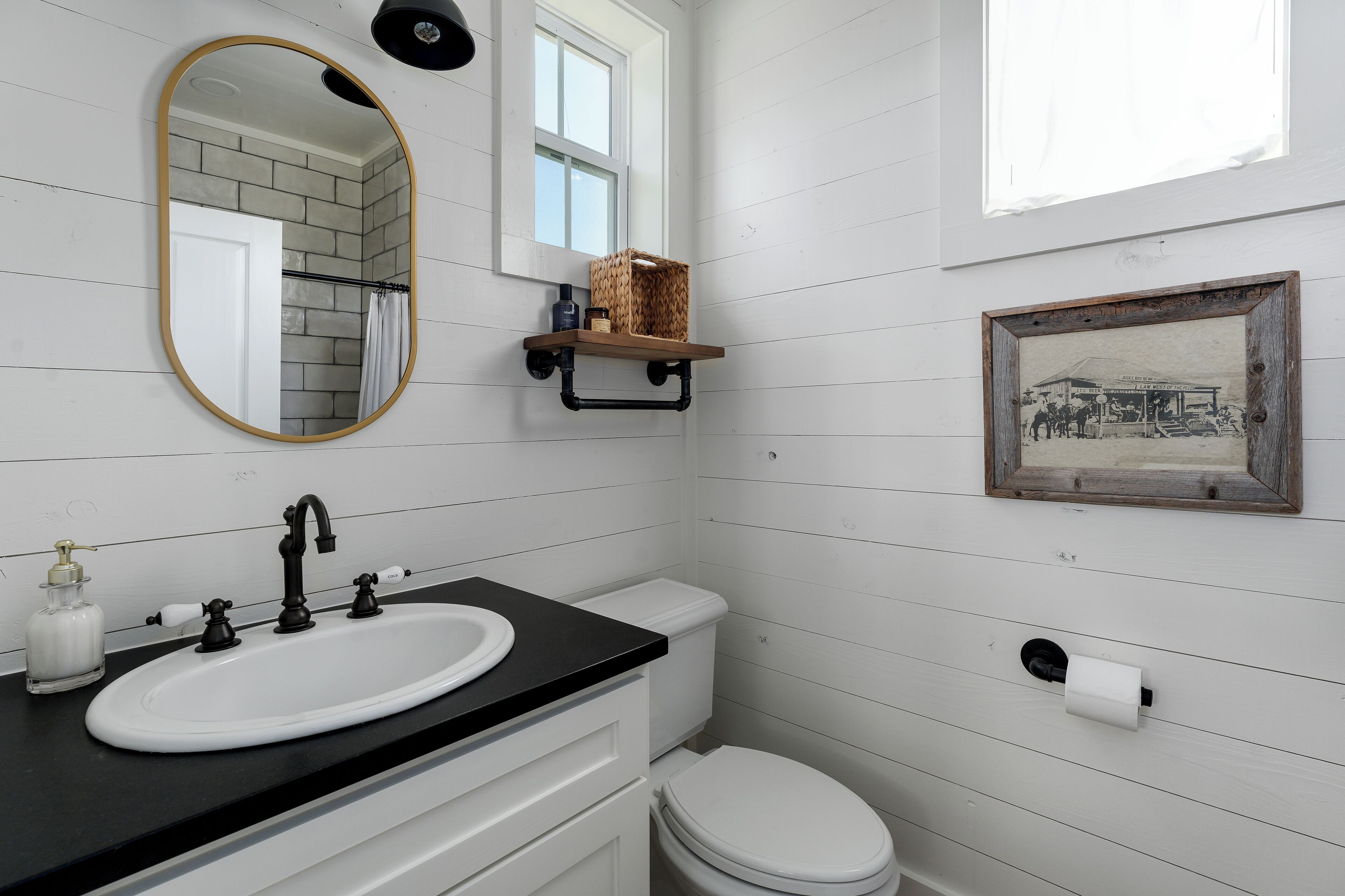 Un baño con lavamanos blanco, grifo negro y un inodoro blanco. Hay un espejo con marco dorado, una ventana y un cuadro en la pared.