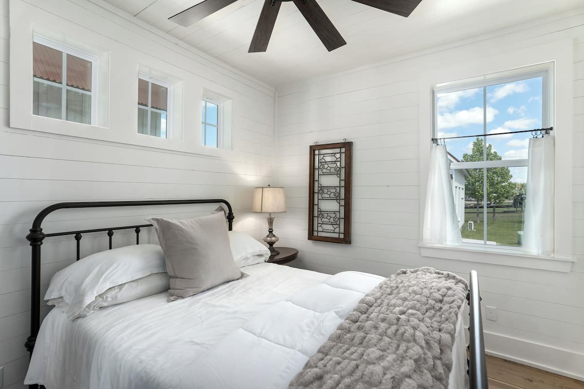 A bedroom with a bed, a nightstand with a lamp, a window with curtains, and a ceiling fan.