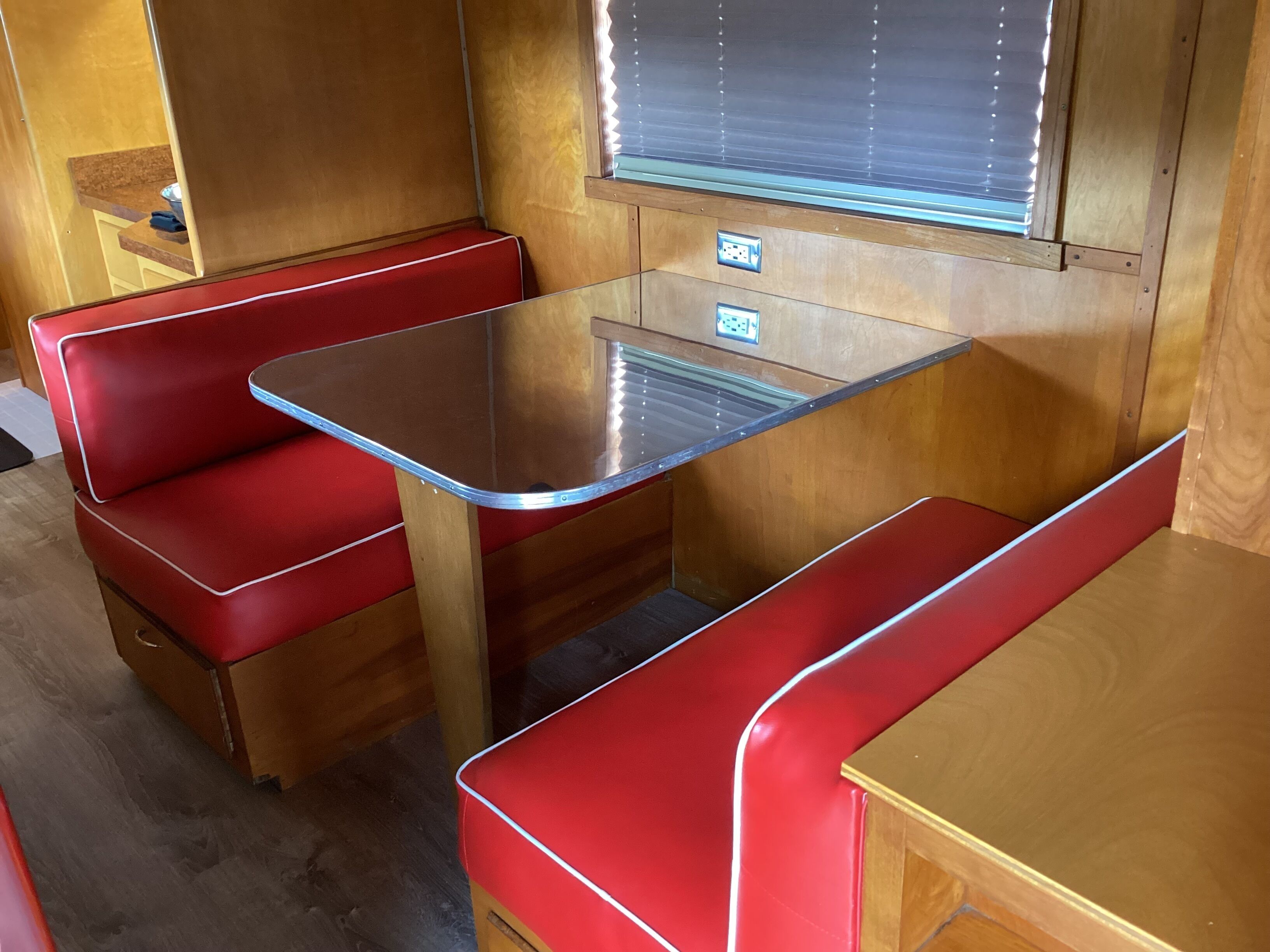 A compact RV interior with a kitchenette area, including a table and two red cushioned seating areas.