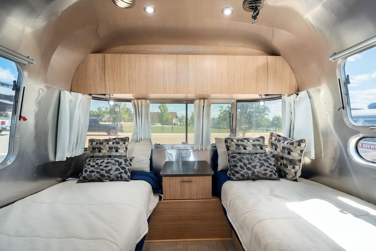 A compact RV interior with two beds, a central table, and large windows offering views of the outdoors.