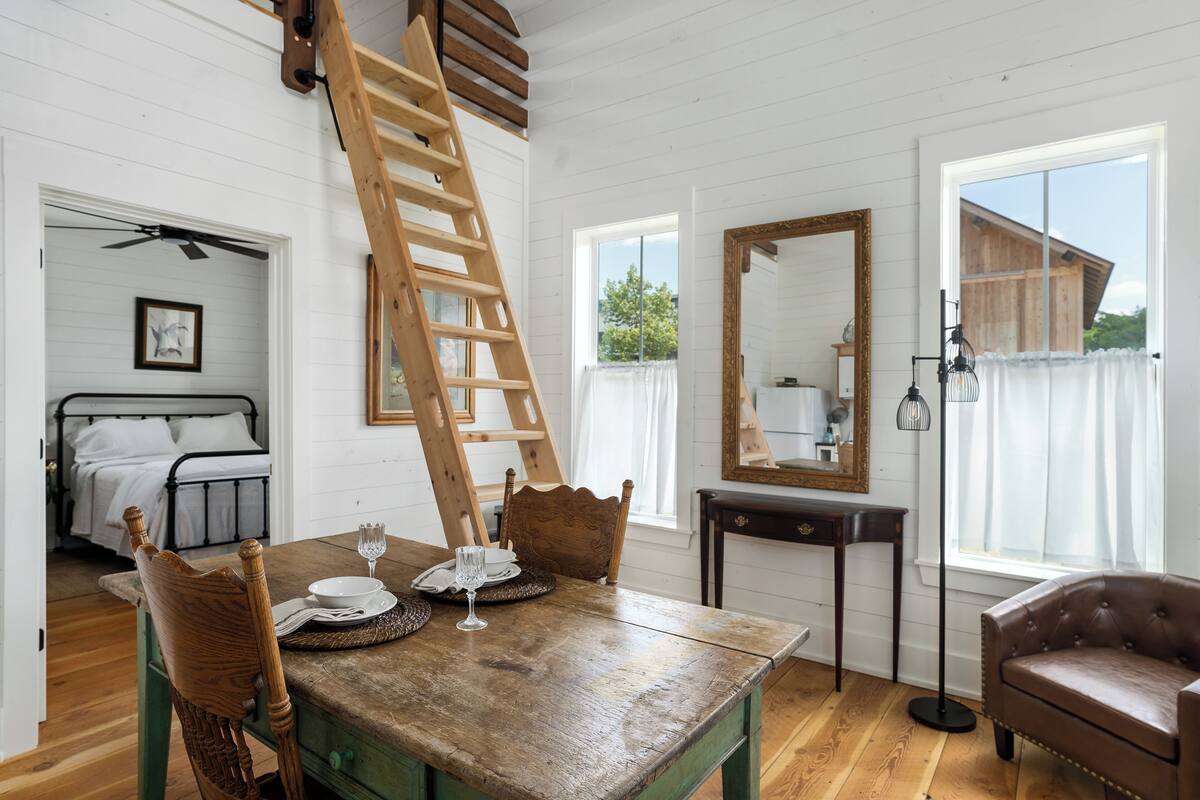 A dining area with a wooden table set for two, a wooden chair, a ladder, a mirror, and a view of a bedroom with a bed and a ceiling fan.
