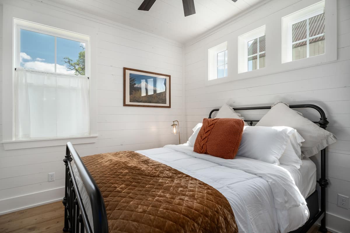 A bedroom with a bed, white walls, wooden floor, and a window with a view of the sky.