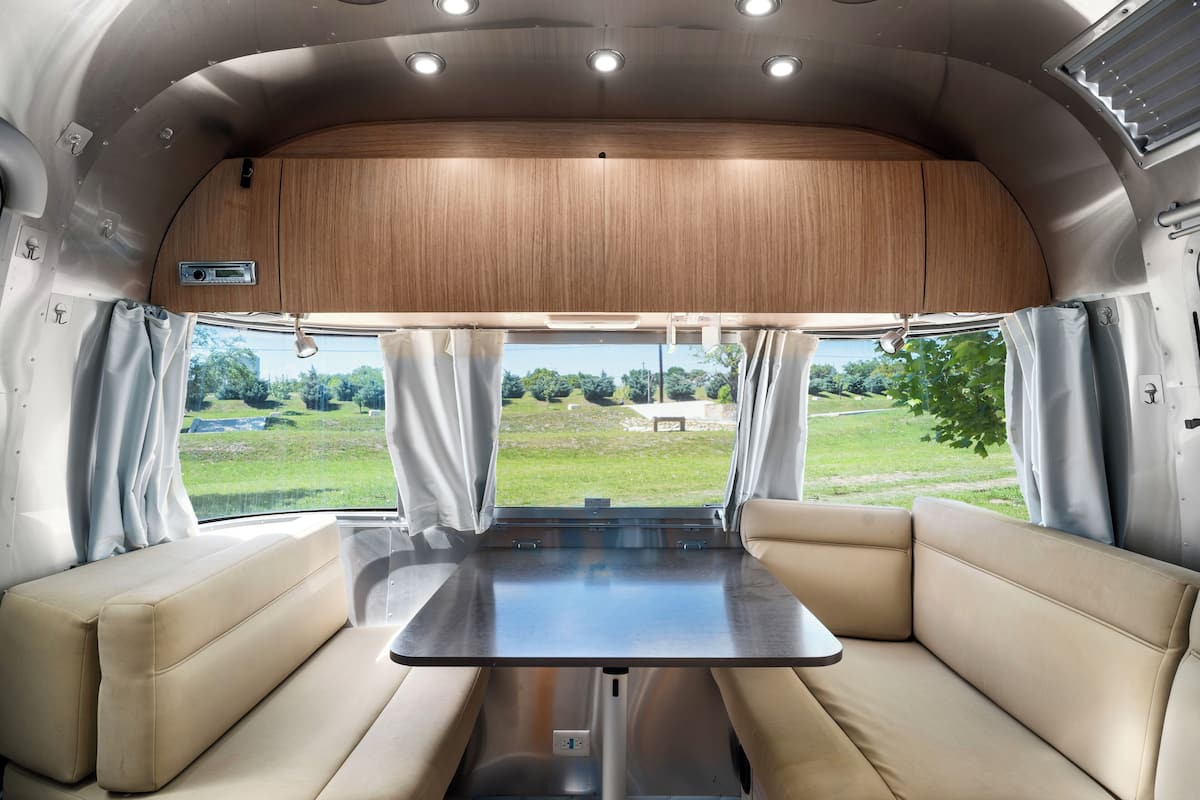 A campervan interior with a central table and cushioned seating, large windows with curtains, and a wooden ceiling.