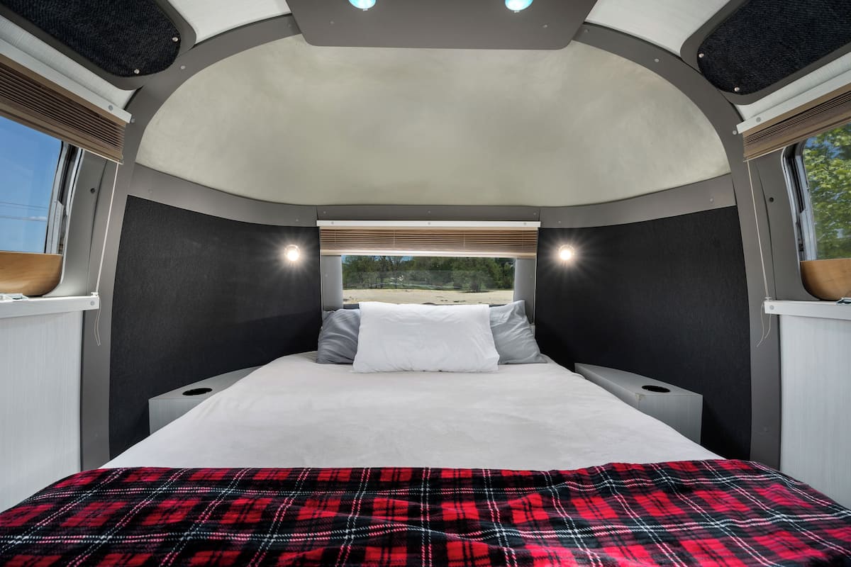 A bed with a red plaid blanket, a window with blinds, and a view of greenery outside.