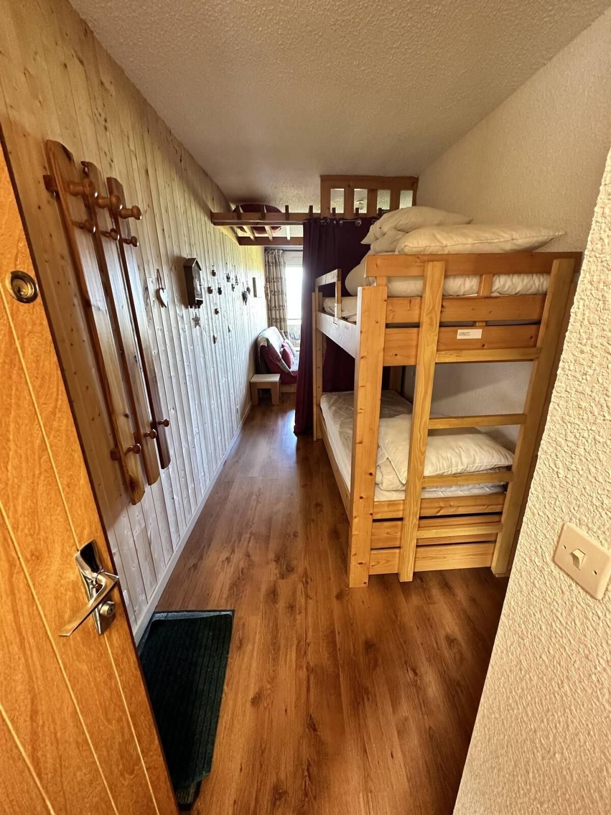 Um quarto com beliches de madeira, uma porta de madeira com puxador de latão e um banquinho de madeira.