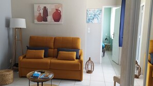 Una sala de estar con un sofá color mostaza, una lámpara, una mesita con revistas y un cuadro de jarrones en la pared.
