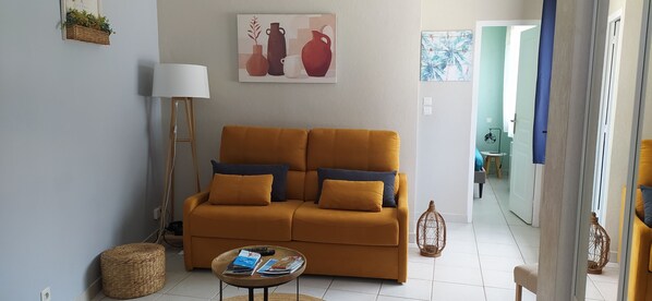 A living room with a mustard sofa, a lamp, a small table with magazines, and a painting of vases on the wall.