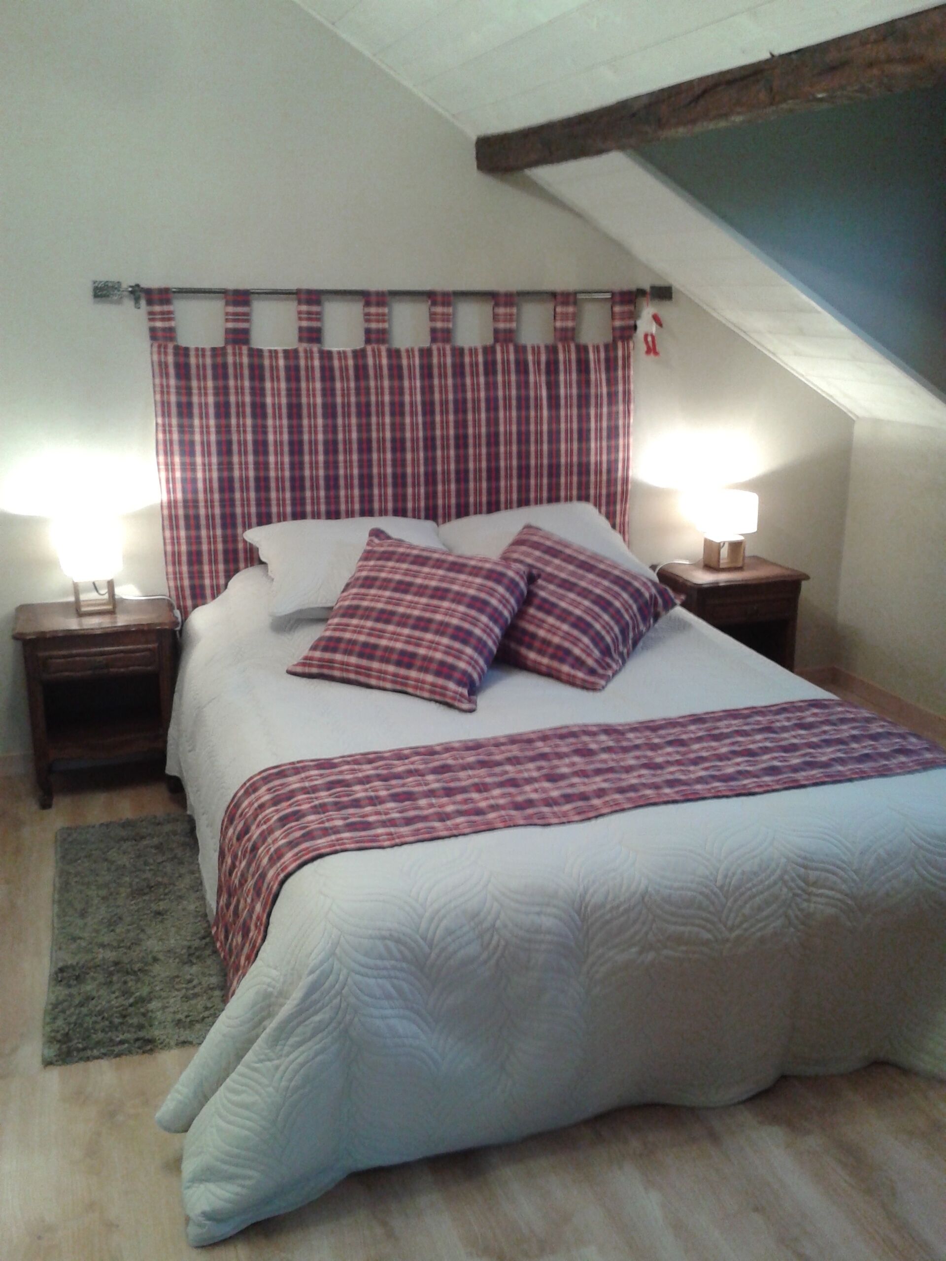 A bedroom with a large bed, plaid curtains, two bedside lamps, a wooden headboard, and a sloped ceiling.