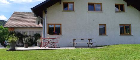 A two-story house with a sloped roof, multiple windows, and a small patio with a table and chairs.
