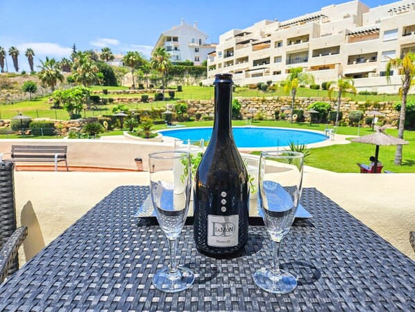 A table with a bottle of wine, two glasses, and a bench in a resort setting with a pool and buildings in the background.