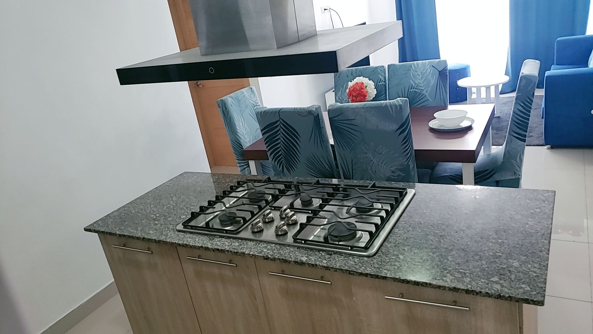 Cozinha com fogão a gás, armários de madeira e área de jantar com cadeiras azuis e uma mesa.