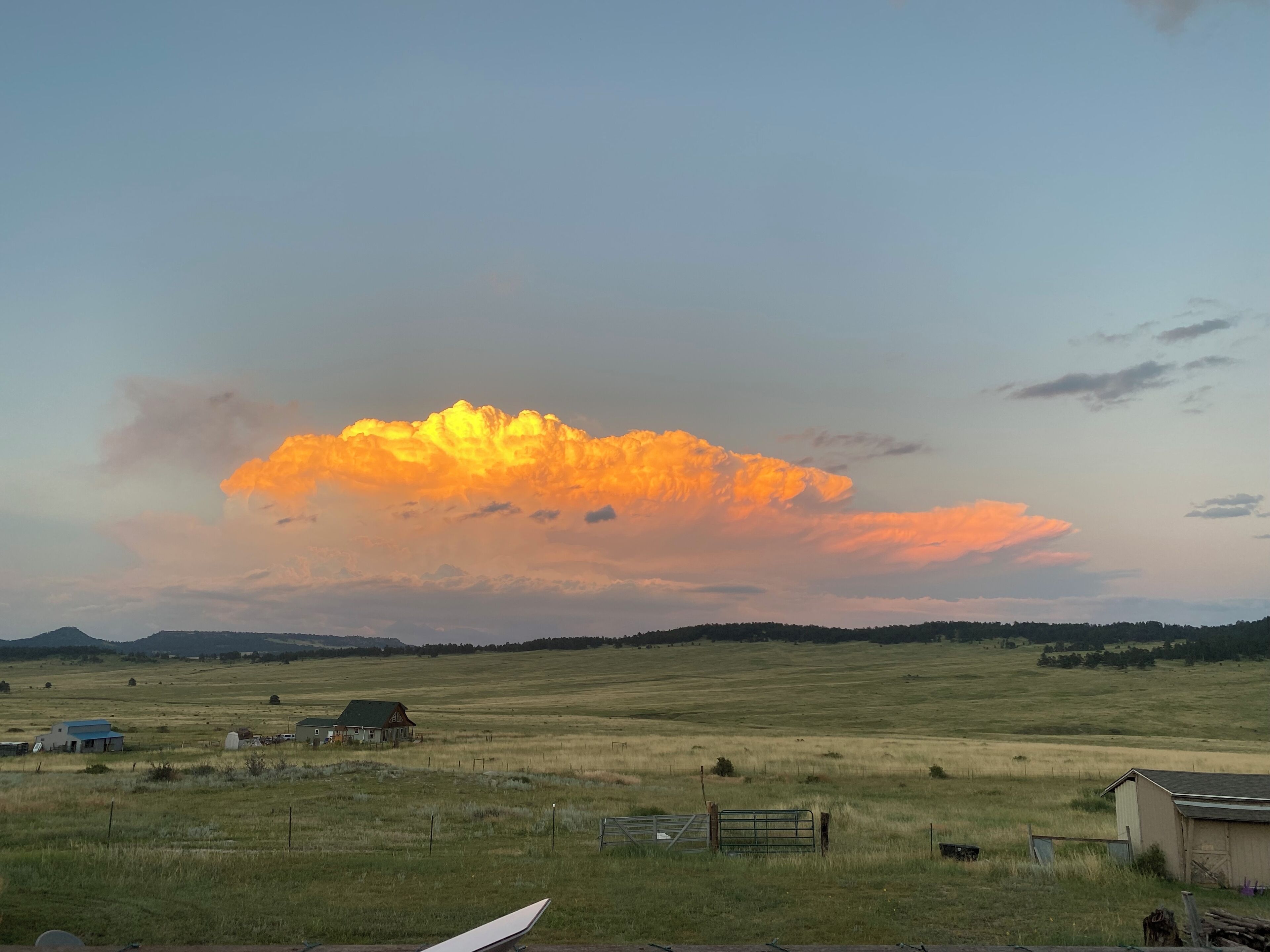 A rural landscape with a large field, a few buildings, and a prominent cloud formation in the sky.