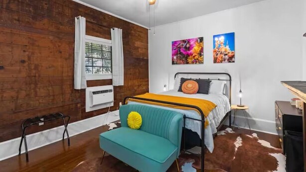 Un dormitorio con pared de madera, una cama con manta amarilla, una silla verde azulada, un ventilador de techo y dos cuadros en la pared.