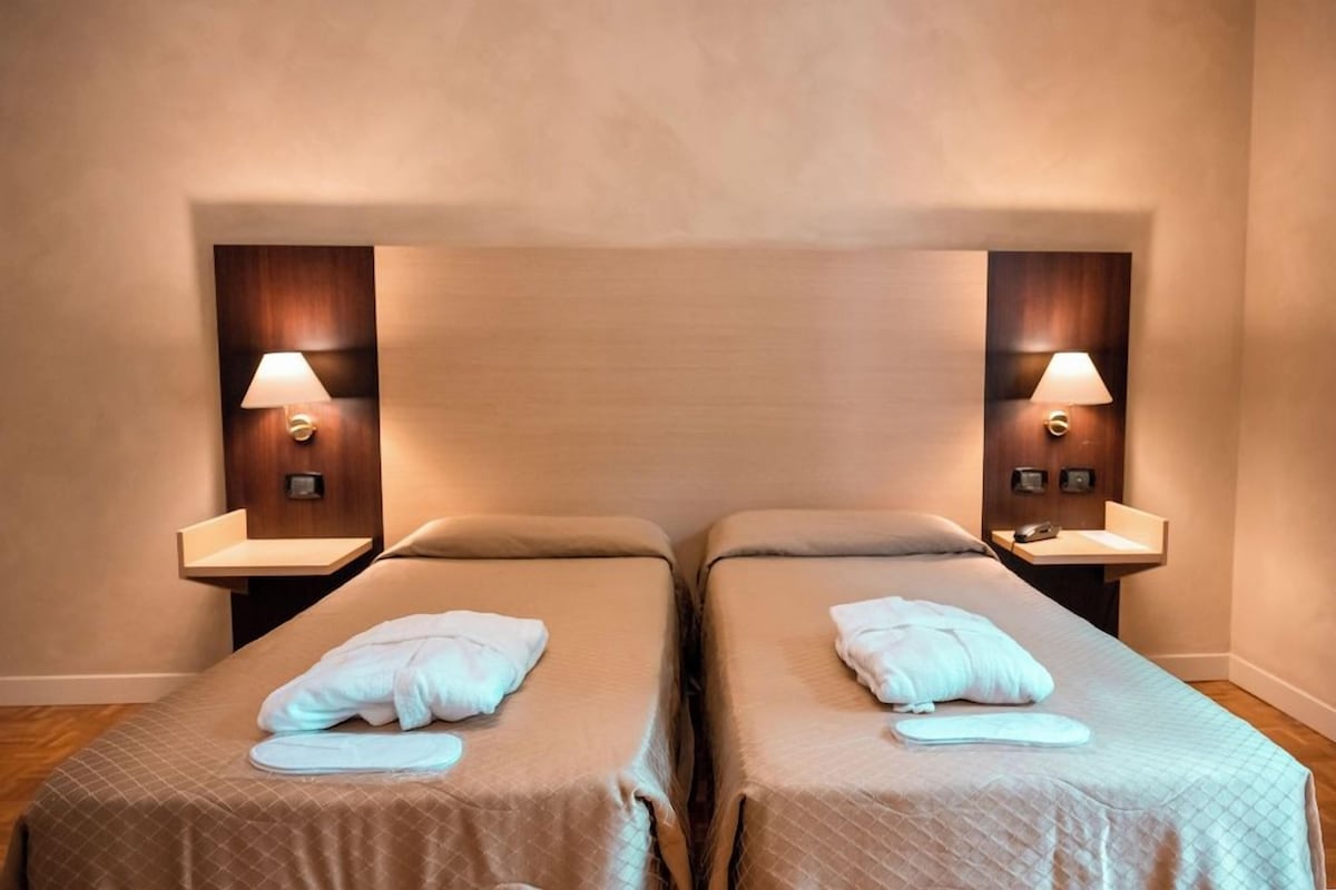 Une chambre d’hôtel avec deux lits simples, chacun équipé d’un oreiller et d’une serviette. On y trouve des tables de chevet avec des lampes et une petite étagère.